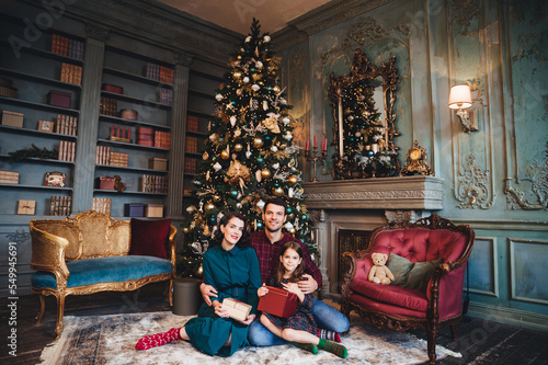Friendly family sits near decorated Christams tree, embrace each other, hold packed presents, enjoy majestic atmospere. Father, mother and daughter near New Year tree in living room. Festive concept © VK Studio