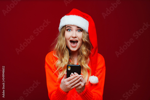 a surprised woman in a Santa Claus hat with smartphone on a red background. 