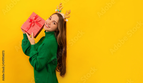 Happy smiling cheerful young woman wearing green knitted sweater with Xmas reindeer antlers, holding pink present gift with bow and looking at camera isolated on yellow background with copy space.