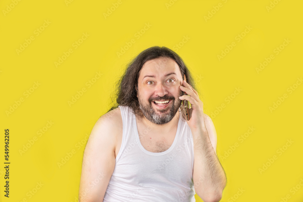 A pleased unshaven man in a white undershirt is talking on a mobile phone with a smile. An unemployed, unkemptly dressed adult from Eastern Europe. A pleased unshaven man in a white undershirt is talking on a mobile phone with a smile. An unemployed, unkemptly dressed adult from Eastern Europe.