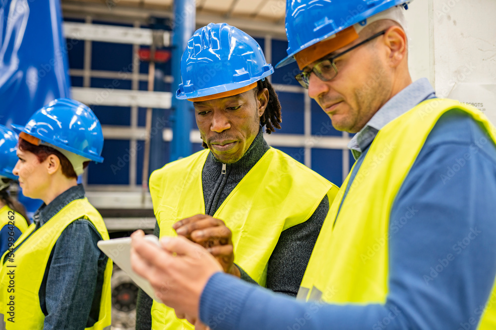Two multiracial workers in safety helmets and reflective vests writing labor actions on a tablet. in a robotic factory. Focus on African American man Two multiracial workers in safety helmets and reflective vests writing labor actions on a tablet. in a robotic factory. Focus on African American man