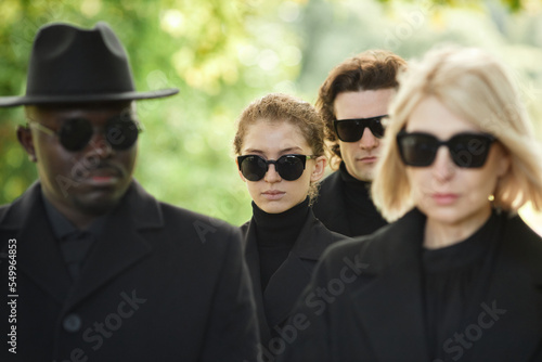 Group of people wearing all black with sunglasses standing in silence at outdoor funeral ceremony