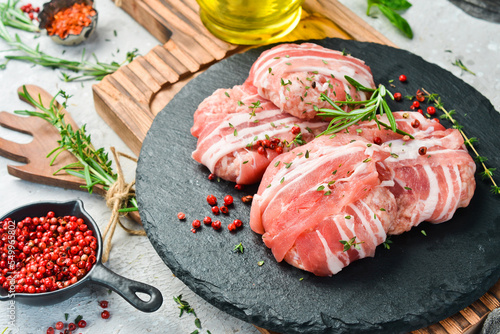 Raw cutlets wrapped in bacon served on a black plate with rosemary and thyme. On a stone background. Top view.