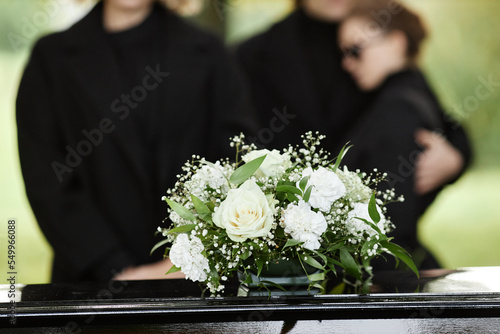 Obraz na plátně Close up of flower bouquet on coffin with people wearing black in background, ou