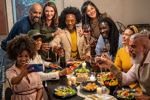 Different people of age and ethnicity eating a vegan dinner. Multiethnic group of friends having fun while sharing a meal in a warm and welcoming house, selfie moment