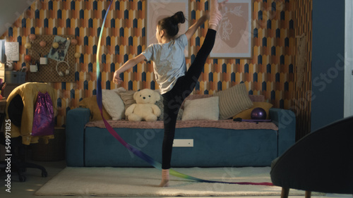 Young asian girl dances with gymnastic ribbon  showing vertical leg-split and spins around herself. Gymnastics training. Sports hobby. Upgrading physical health on quarantine. Slow motion.