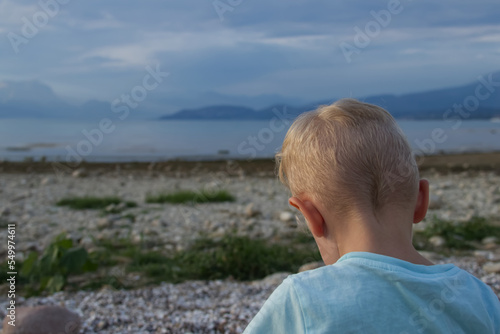 A little boy sits with his back on the shore of the lake against the backdrop of blue mountains 