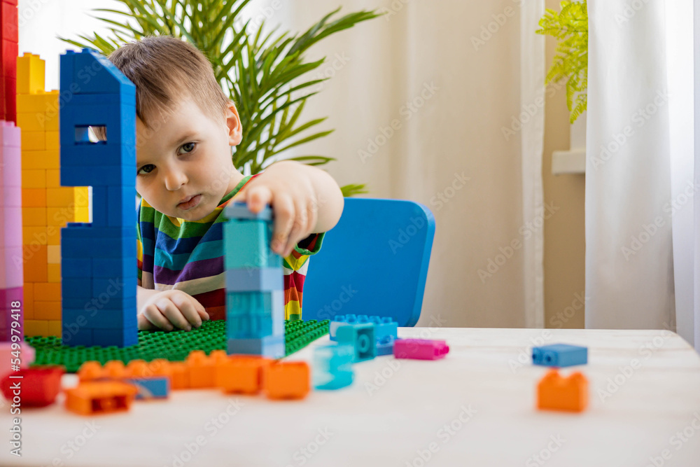 Confident male baby constructing tower with multicolored plastic pieces assembling house on table Confident male baby constructing tower with multicolored plastic pieces assembling house on table