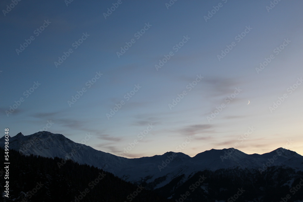 Sunset over Swiss alpine mountains. Silhouette of a mountain range against sky at sunset with gradient from indigo to light pale yellow. Snow on peaks Sunset over Swiss alpine mountains. Silhouette of a mountain range against sky at sunset with gradient from indigo to light pale yellow. Snow on peaks