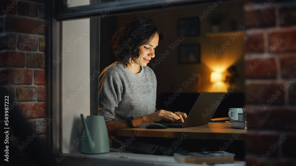 Smiling Hispanic Woman at Home Using Laptop During Warm Cozy Evening. Professional Freelancer Working Remotely From Stylish Home. Shot From Outside Into the Window of Apartment.