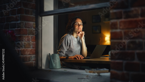 Smiling Hispanic Woman at Home Using Laptop on the Warm Cozy Evening and Looking Thoughtfully. Professional Freelancer Working Remotely From Home. Shot From Outside Into the Window of Apartment.