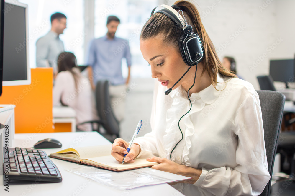 Beautiful young lady working in office. Customer support worker taking notes while talking with client via headset at office desk. Beautiful young lady working in office. Customer support worker taking notes while talking with client via headset at office desk.