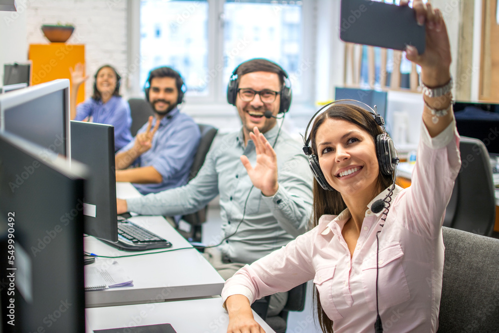 Obraz premium Beautiful woman customer support operator taking a selfie photo of her team colleagues in a call center.
