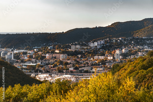 View from the height of the seaside town on a sunny autumn day. The mountains surrounding the town are covered with autumn vegetation under a blue cloudy sky.