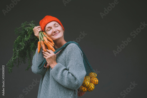 Smiling woman with eyes closed holding carrots and oranges in front of gray wall