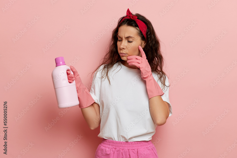 a young housewife stands against a pink wall, she looks thoughtfully at a bottle of cleaning agent in her hand a young housewife stands against a pink wall, she looks thoughtfully at a bottle of cleaning agent in her hand