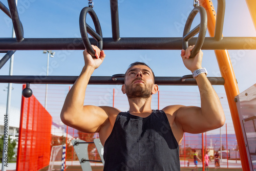 Man in a black t-shirt pulls up on gymnastic rings outdoors, exercising on the sports ground.