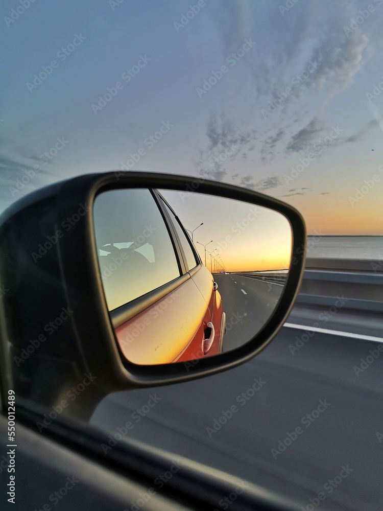 Reflection of road and sunset in the side mirror of the orange car. Concept of road trip and traveling by car. 