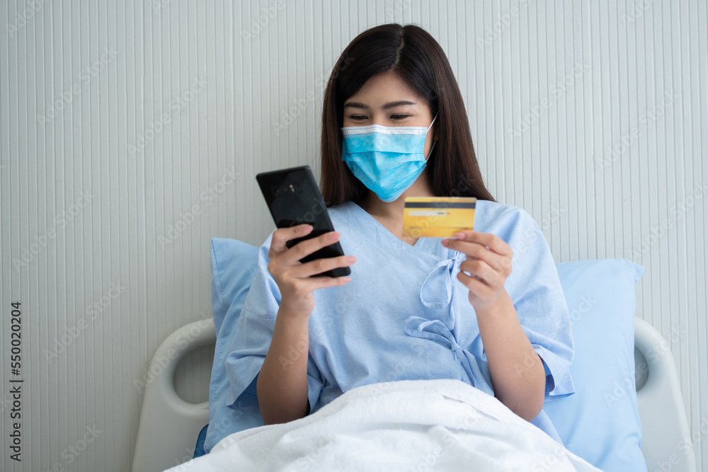 Happy Asian woman wearing a medical mask and holding mock up credit /insurance card and smartphone in a hospital bed for Check health insurance eligibility. Insurance policy by bank, payment medical Happy Asian woman wearing a medical mask and holding mock up credit /insurance card and smartphone in a hospital bed for Check health insurance eligibility. Insurance policy by bank, payment medical