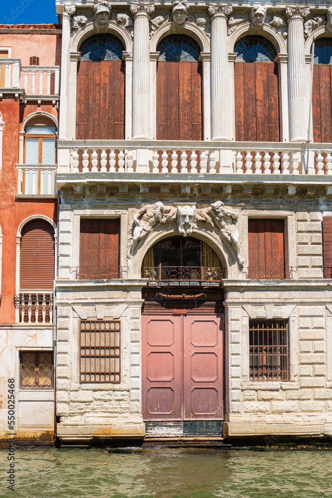 Facade of a vintage building on a Venetian canal. Wooden shutters closed the windows on the wall of an old house at the water's edge on a street in the center of Venice, Italian aesthetics