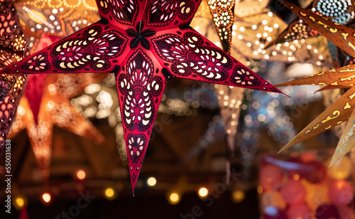 Many colorful stars made of colorful cardboard are illuminated from the inside. The stars hang from the ceiling. The background is lit up for Christmas.