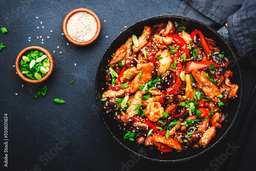 Stir fry with turkey fillet, paprika, mushrooms, green chives and sesame seeds in frying pan. Asian cuisine dish. Black stone kitchen table background, top view
