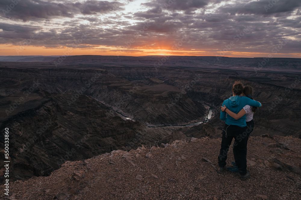 Dramatischer Sonnenuntergang - Frau und Tochter blicken umarmt auf eine Schleife des Fish Rivers bei Sonnenuntergang, wie er sich durch den Fish River Canyon windet (Namibia)