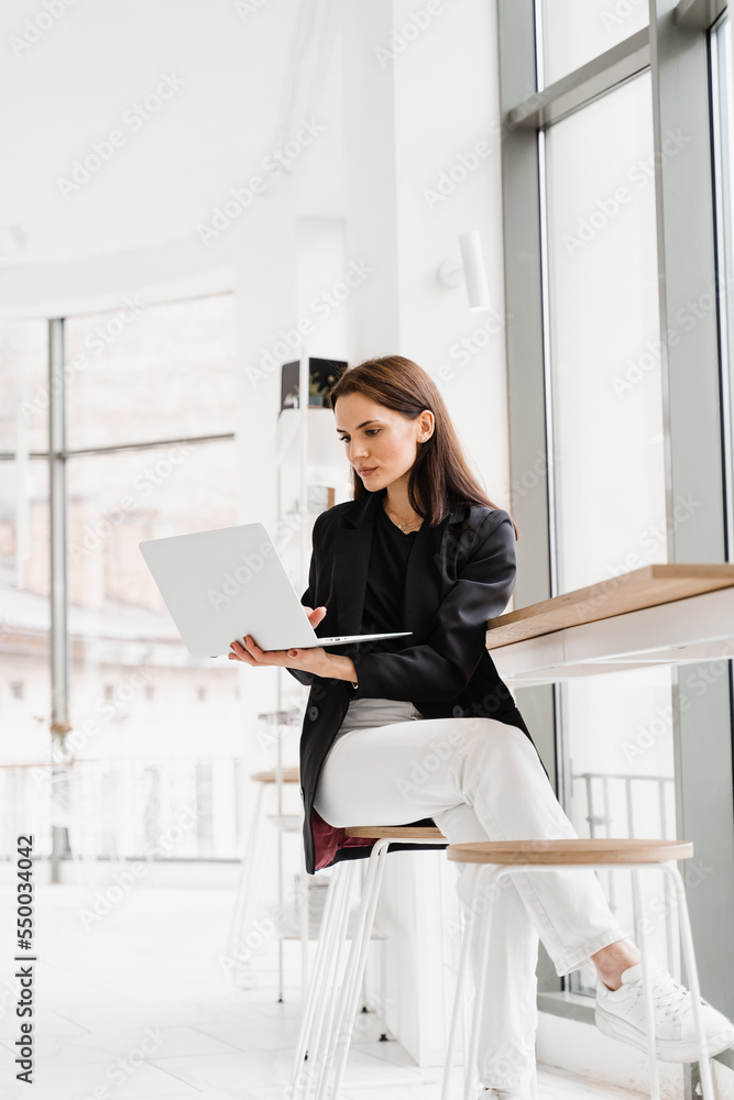 Confident young business woman working online with team on laptop on startup project. Attractive girl programmer with laptop is working online in white cafe.