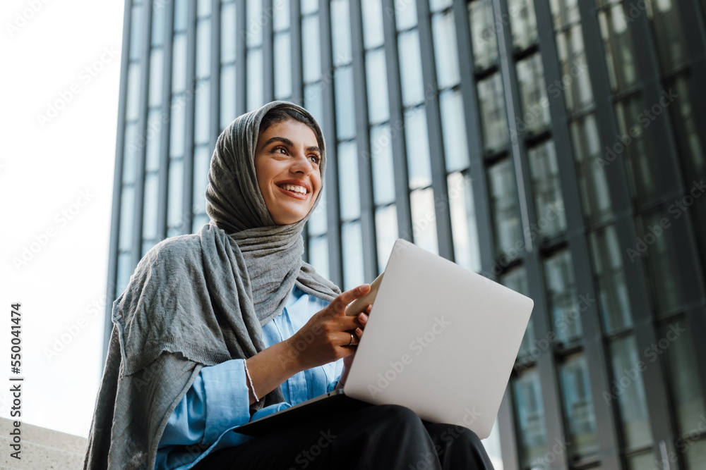 Young muslim woman wearing headscarf using cellphone and laptop outdoor - obrazy, fototapety, plakaty 