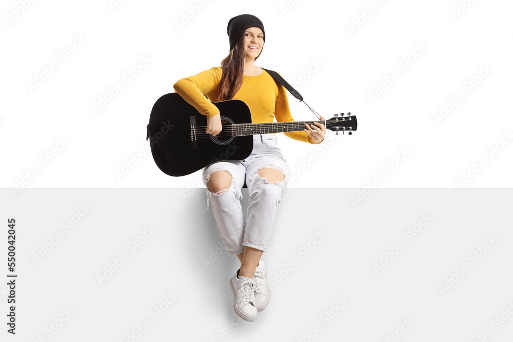 Young female musician playing an acoustic guitar and sitting on a blank panel Young female musician playing an acoustic guitar and sitting on a blank panel