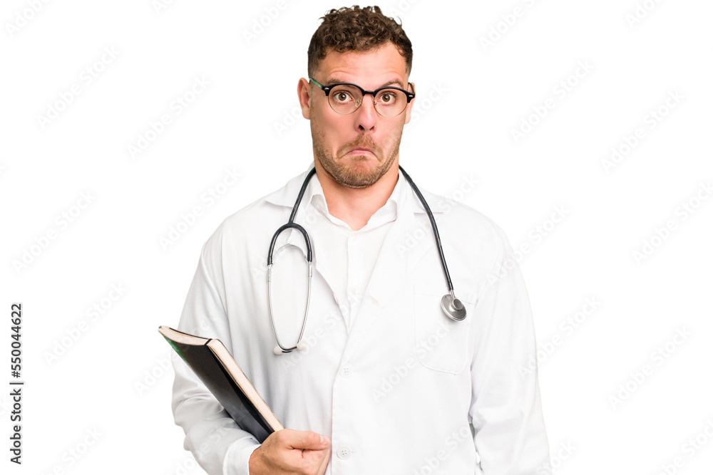 Young doctor caucasian man holding a book isolated shrugs shoulders and open eyes confused. Young doctor caucasian man holding a book isolated shrugs shoulders and open eyes confused.