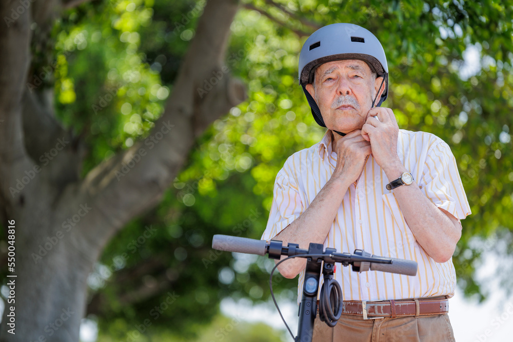 © alex.pin - uomo anziano si sistema il  caschetto vicino al suo monopattino elettrico in una pista ciclabile vicino a un parco cittadino .
