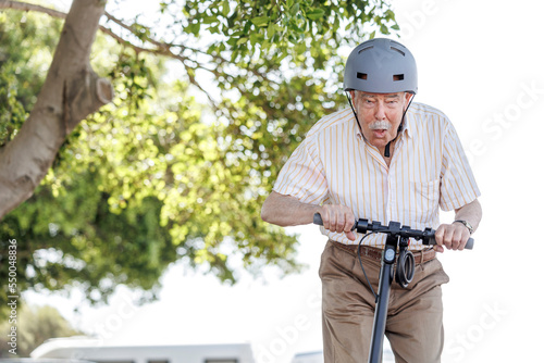 Konstfotografi uomo anziano con il caschetto , monopattino elettrico corre con espressione impegnata in una pista ciclabile vicino a un parco cittadino
