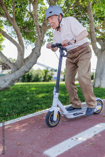uomo anziano con il caschetto , monopattino elettrico corre con espressione impegnata in una pista ciclabile vicino a un parco cittadino .