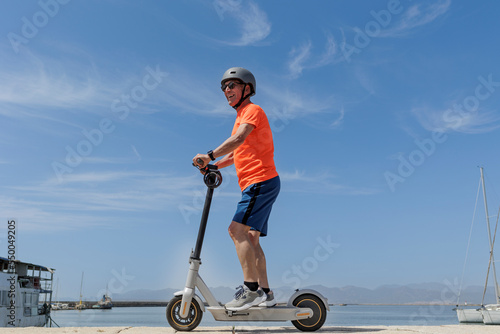 Uomo anziano con una maglia arancione e caschetto protettivo , corre sul suo monopattino elettrico  di fronte al porto turistico ., isolato su sfondo cielo .  photo