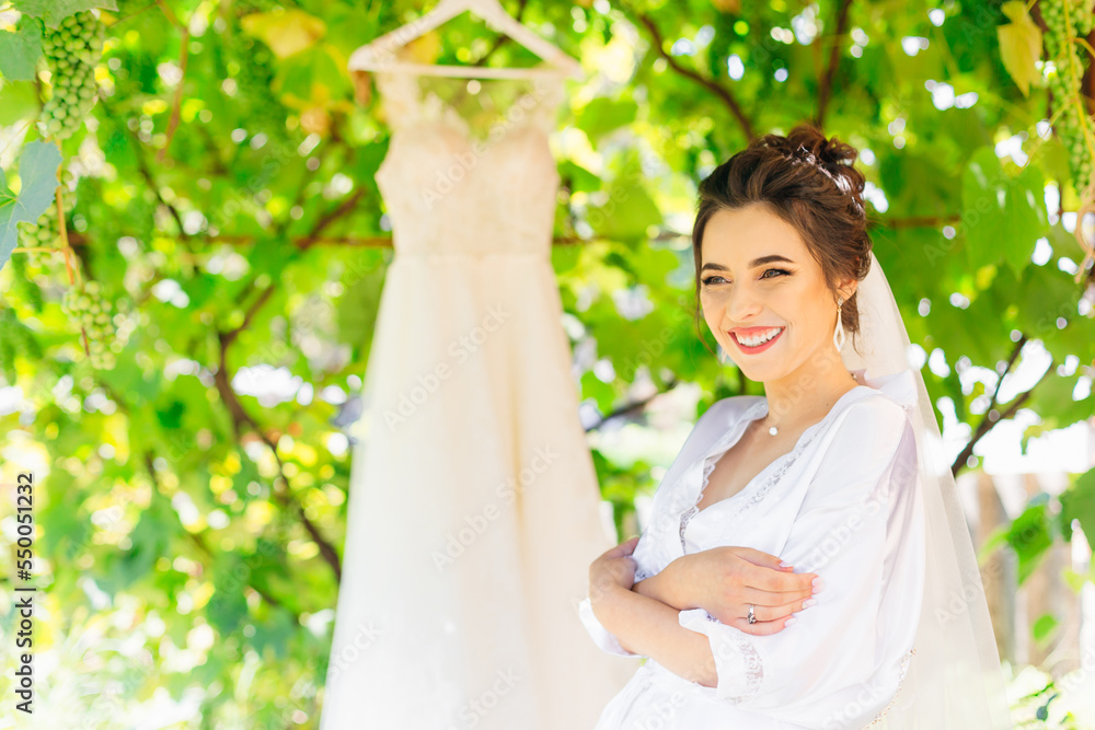Stunning woman in lingerie smiles sincerely and looks away while posing outdoors. The bride enjoys every moment of the big day. The wedding dress hangs on a tree behind. Stunning woman in lingerie smiles sincerely and looks away while posing outdoors. The bride enjoys every moment of the big day. The wedding dress hangs on a tree behind.