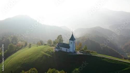 Aerial view of small church on the hill and low clouds at sunrise in autumn. Slovenia. Top view from drone of beautiful chapel on mountain in fog, green meadows, trees, sky at dawn in fall. Landscape photo