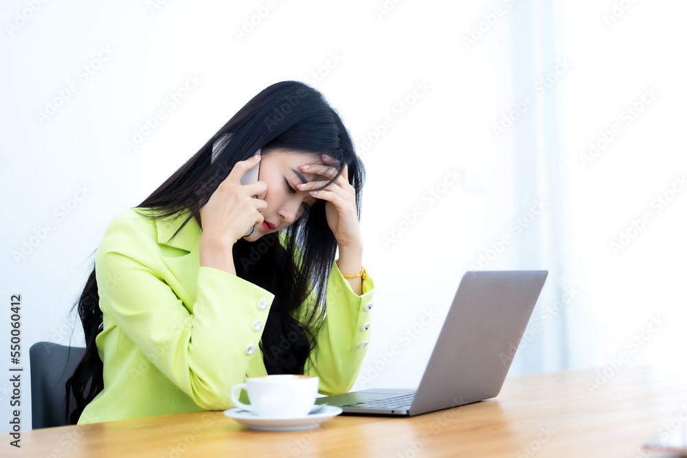 Young business woman wears green suit using laptop computer and mobile phone. Beautiful woman talking on the phone feeling sad and moody. Business woman looking irritable while talking on the phone.