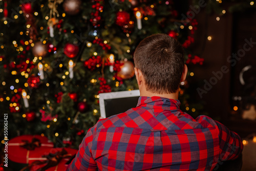 Young handsome man in winter shirt is working at white laptop at home near the Christmas tree. View from the back.
