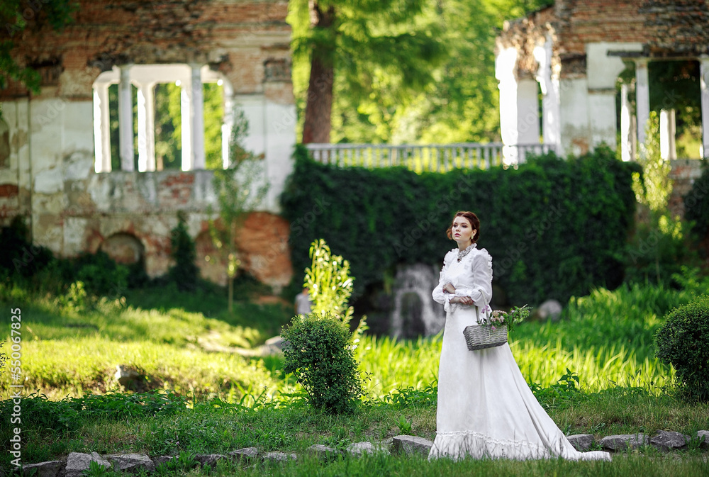 Fototapeta premium Beautiful girl in a vintage long white dress, and a hairstyle in the English style in a summer park, against the background of columns, warm summer