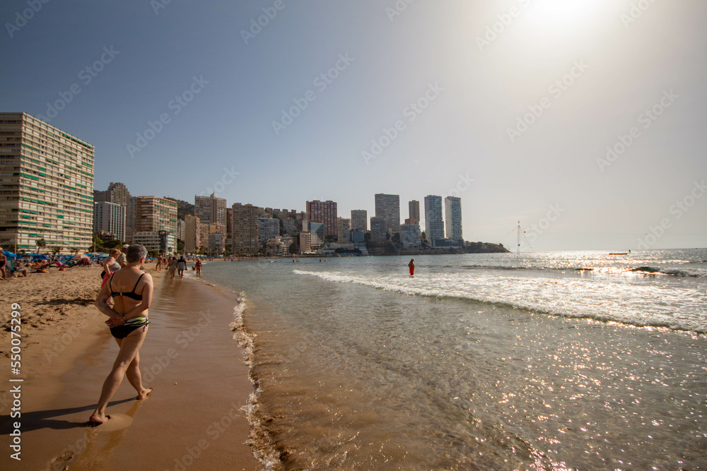 Personas caminando por la orilla de la playa de Benidorm en bañador bajo un precioso día soleado de otoño con laos grandes hoteles de fondo. Personas caminando por la orilla de la playa de Benidorm en bañador bajo un precioso día soleado de otoño con laos grandes hoteles de fondo.