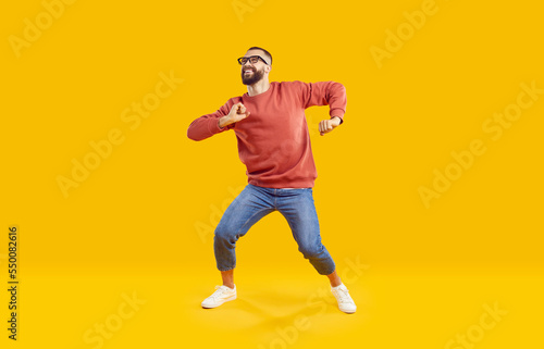 Cheerful active man enjoying life, rejoicing, dancing and having fun on orange background. Cool stylish Caucasian bearded man in jeans and sweatshirt dancing in good mood. Full length. Web Aner.