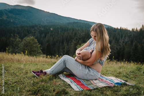 Family hiking and feeding infants everywhere outdoors. Mother feeding breast newborn baby sitting on blanket on grass in the field. World Breastfeeding Week. Mom hugs child and travels in mountains.