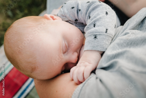 Breastfeeding baby. Mother breast feeding newborn girl sitting on grass in park. Concept of lactation infant, postpartum period, natural motherhood. Mom hug daughter on nature. Babywearing. Closeup.