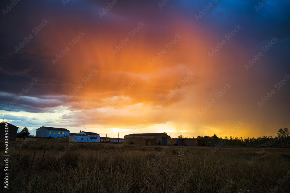Fototapeta premium small houses of village with orange sky during storm rains at sunset