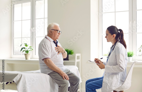 Agitated elderly man with glasses holds his hand on his chest, talking to a doctor in white coat, sitting on medical couch. Beautiful young female doctor supports unhealthy elderly man on consultation