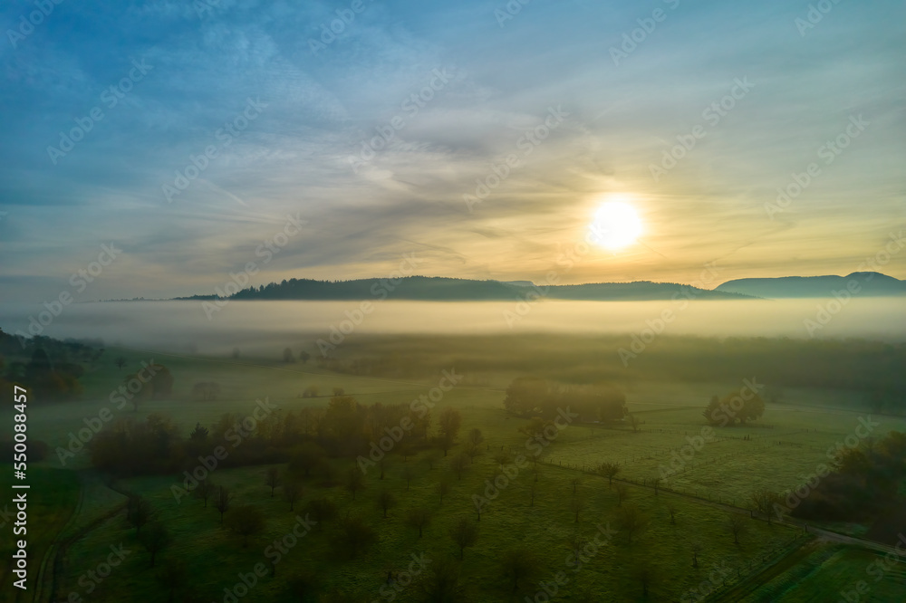 Aerial view of fog landscape at sunrise. Hill range behind agricultural fields in autumn. Blue sky for copy space. Germany, Nurtingen, Tiefenbachtal.