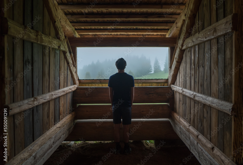 Young lonely teenage boy standing near the wooden forest house window and enjoying the morning spruce foggy forest. Traveling and tourism at fall time. Young lonely teenage boy standing near the wooden forest house window and enjoying the morning spruce foggy forest. Traveling and tourism at fall time.