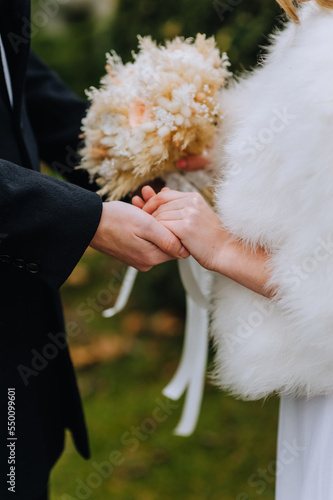 A stylish groom in a black coat and a bride in a white fur coat with a bouquet of wild flowers and reeds gently hold hands. Wedding photography, portrait, ceremony.