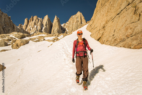 MT WHITNEY, MOUNTAINEER'S ROUTE, LONE PINE, CA, USA. A 30 year-old woman in mountaineering clothes smiles as she descends a steep snow field with jagged granite peaks rising behind photo